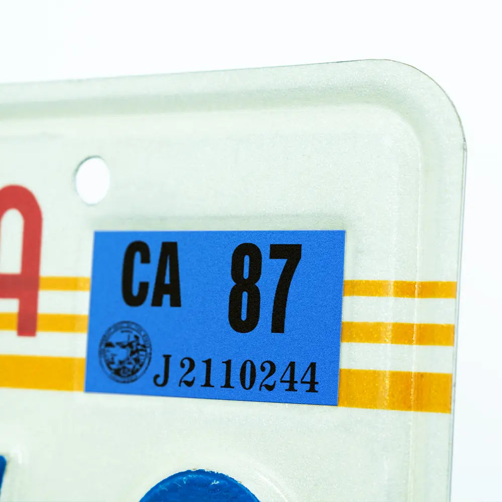 Close-up of a blue 1987 California registration sticker mounted on a white California license plate with yellow reflective stripes.